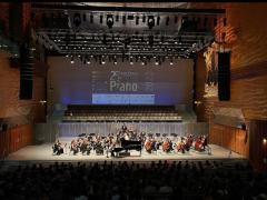 Foto do palco do 25o Concurso Internacional de Piano Santa Cecília. Na imagem, uma orquestra se apresenta para um público em uma sala de concerto. Ao centro do palco, um piano grande e preto. Ao fundo, no telão, aparece o cartaz de divulgação do concurso. A iluminação é direcionada ao palco e as paredes do ambiente têm tons de marrom e laranja. 