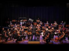 Foto de uma apresentação da Orquestra Parassinfônica de São Paulo. Pessoas, a maioria sentadas, vestidas de preto, tocam instrumentos como violoncelo, viola e violino em um palco de madeira. No centro, de costas para a câmera, um regente, com terno preto, movimenta os braços. A iluminação se concentra nos musicistas, ao redor deles há pouca luz. No fundo, uma cortina azul escuro. 