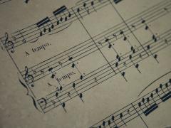 Foto de uma folha de partitura impressa. O papel é levemente amarelado, com algumas manchas. Sobre ele, parte das linhas da partitura, divididas em blocos de cinco linhas cada, que se alternam, no sentido horizontal, com espaços em branco. Sobre as linhas há diferentes notações musicais e em dois dos espaços em branco aparecem os dizeres “a tempo”. Todos os elementos impressos são da cor preta. 