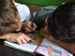 Foto de duas crianças, uma de pele clara, cabelos castanhos e camiseta branca, e outra de pele parda, cabelos pretos, colete verde e camiseta branca, debruçadas sobre um livro com texto e ilustrações coloridas de pessoas. A criança parda escreve sobre o livro enquanto a outra aponta para um dos desenhos. Ao fundo, uma parede verde.