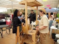 Foto de uma feira em um pátio coberto. Em primeiro plano, estão duas pessoas conversando em pé separadas por uma banca de madeira com utensílios para fazer café. A pessoa da esquerda é negra, possui cabelos médios, castanhos e cacheados. Ela usa óculos, blazer preto e camisa branca. Do outro lado da banca, há uma pessoa branca, de cabelos curtos e castanhos. Ela usa chapéu cor de palha, camisa branca e calça bege. Ao fundo, há mais bancas, com produtos e pessoas circulando, cadeiras de plástico e plantas. 