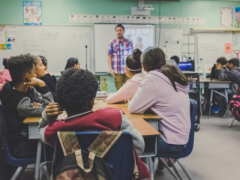 Fotografia de uma sala de aula com parede verde, piso cinza, quadro branco e cartazes. Estudantes  estão sentados em grupos em carteiras dispostas na sala com a atenção voltada ao professor, um homem branco que veste camisa xadrez e está à frente do quadro branco. 