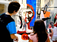 Uma criança de cabelos compridos e blusa Rosa e uma joem de cabelos curtos e blusa preta estão em pé, de costas, observando uma obra de arte, o desenho do busto de um homem, feito em materiais diversos, nas cores preto, vermelho azul e transparência. Ao fundo, prédios e a calçada mostram que elas estão em um espaço aberto da cidade de São Paulo