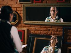 Foto de uma cena do documentário de Sérgio, onde ele está diante de vários espelhos. Sérgio é um homem indígena, de cabelos grisalhos e veste uma camisa social branca sob um colete colorido. Está em frente a cinco espelhos, pendurados numa parede de tijolos laranjas. É possível ver o seu reflexo em dois desses espelhos.