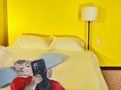 Ilustração de uma cama de casal a frente de uma parede amarela vibrante. Nela há dois travesseiros e a pintura de um macaco deitado em um cobertor azul vestindo um vestido vermelho e segurando um celular com as duas patas. Ao lado da cama há um abajur grande e branco, de haste preta e fina e ao seu lado há um interruptor. O chão é texturizado em vermelho e o rodapé marrom. 