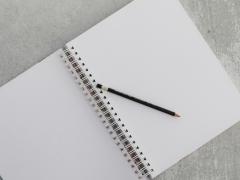Foto de um caderno com espiral aberto e sem pauta, sobre o qual está apoiado um lápis preto com borracha em uma das pontas. O caderno está sobre um fundo cinza.