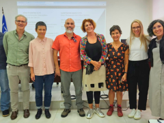 Foto de sete pessoas de pé em uma auditório, lado a lado, posando. Ao fundo, uma parede branca, com uma projeção, e as bandeiras de São Paulo, Do brasil e da USP, ao fundo, do lado esquerdo.