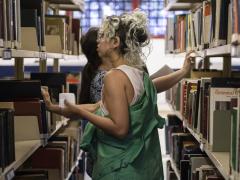 Foto de duas mulheres entre duas prateleiras de livros de uma biblioteca. Em primeiro plano, virada para a esquerda é branca, tem cabelo ondulado e castanho com mechas loiras, usa brinco de argolas, uma regata branca e uma jaqueta verde. Ela segura um pequeno papel em uma das mãos e a outra apoia na prateleira de livros. A de trás, a qual se pode ver parcialmente, está virada para a direita,  é branca, usa uma camiseta estampada branca e azul e tem cabelos castanhos, lisos e longos que estão presos.