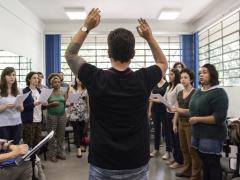 Foto de um grupo de coral em sala de aula sendo regido por um homem de costas para a foto. O regente está de braços erguidos, juntando o indicador e o polegar nas duas mãos. Ele é branco, tem cabelos castanhos curtos, usa uma camiseta preta, um relógio no braço esquerdo, que é parcialmente coberto por tatuagens. À frente do regente há um grupo cantando, tendo fileiras femininas à frente e masculinas ao fundo. Do lado esquerdo da imagem, uma pessoa sentada faz anotações em um papel sobre estante de partitura