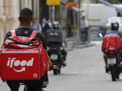 Foto de entregadores de aplicativo em uma rua. No lado esquerdo, uma pessoa negra, de cabelo raspado e de costas, pedala uma bicicleta. Ela usa camiseta preta e uma grande mochila vermelha em formato quadrado, a bag, com o logo do Ifood. Em segundo plano, um pouco desfocados, duas pessoas em motos: a do lado direito tem a pele parda, usa capacete e bermuda pretos, regata azul e bag  vermelha. A pessoa à  esquerda usa capacete e calça pretos e tem o resto do corpo coberto pela bag, que é preta.