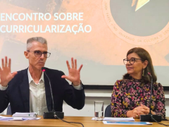 Foto em plano médio. À esquerda, homem branco, de cabelos curtos e grisalhos, usando óculos de grau de haste escura e uma camisa branca quadriculada e um blazer preto. Ele está sentado, ergue as duas mãos para cima, na altura do ombro, enquanto fala ao microfone. Ele é observado por uma mulher que está sentada à direita. Ela é uma mulher branca, de cabelos castanhos, lisos e na altura dos ombros. Ela usa óculos de haste escura e arredondada, brincos dourados, e um vestido preto de manga longa com estampa.