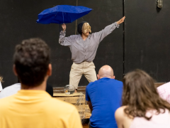 Foto de uma performance em que uma pessoa está com uma máscara dourada de rosto triste. Com a mão direita ela segura um guarda-chuva azul e estende a esquerda na diagonal alta. Ela usa camisa cinza, calça bege e se apresenta diante de uma pequena platéia de costas para a foto. O cenário é preto.