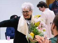 Foto de Maria Immacolata Vassallo de Lopes, uma mulher idosa, de pele e cabelos brancos lisos e curtos, óculos de armação escura e batom vermelho. Ela usa um casaco preto de gola creme e segura um buquê de flores em tons de rosa e amarelo. Ela sorri e acena enquanto algumas pessoas batem palmas no canto inferior direito da imagem. Ao fundo, algumas pessoas estão de pé e sorriem. 