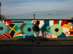 Foto de uma pessoa caminhando em uma calçada, tendo ao fundo um muro grafitado. O grafite é composto por círculos grandes azuis ou verdes que têm em seu centro círculos pretos. Em volta deles, há formas coloridas como retalhos de bandeiras de países. A pessoa  usa um moletom preto com capuz cobrindo o rosto, calça jeans, tênis, e carrega uma mochila e um objeto de metal. Atrás do muro, há o topo de duas árvores e um poste, sob um céu azul.