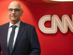 Foto de um homem branco sorrindo. Ele veste terno e gravata azuis, óculos de armação quadrada e preta. Ao fundo, uma parede vermelha e um letreiro com o texto “CNN”.