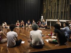 Foto de um grupo de aproximadamente 15 pessoas jovens sentadas em roda no chão de madeira de um grande palco. Ao fundo há uma parede alta e preta e, na lateral direita, uma mesa retangular comprida e branca e uma parede com várias cordas de manobra de teatro na vertical.