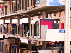 Foto de três prateleiras de uma estante de livros de uma biblioteca, posicionada em diagonal em relação ao enquadramento da imagem. A estrutura da estante é bege e os livros, que possuem etiquetas brancas na lombada, são de diversas cores e tamanhos. Ao fundo, há uma parede branca. 