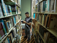 Foto de um rapaz folheando um livro em uma biblioteca.  Ele usa óculos, tem cabelos curtos e pretos, veste roupa esportiva e está entre dois corredores de estantes cheias de livros.