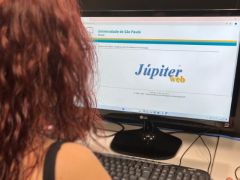 Fotografia de uma mulher olhando a tela de um computador. A mulher está de costas para a câmera, tem pele branca e cabelo vermelho e observa a tela do computador onde é há uma logo escrita “Júpiter web”. 