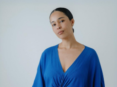 Na foto, uma mulher negra com cabelos pretos presos em um coque baixo. Ela veste blusa azul com decote V e usa brincos. O fundo é cinza.