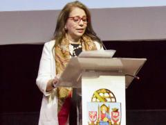 Foto de uma mulher branca, que discursa, de pé, atrás de um púlpito. Ela tem cabelos longos e loiros e usa óculos com armação vermelha, veste um blazer branco e tem um lenço estampado em volta do pescoço. Ela olha ligeiramente para baixo, para ler um papel a sua frente. Atrás dela, um fundo preto e cinza. 