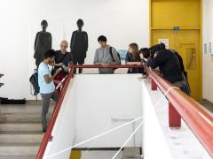 Foto de um grupo de sete pessoas que conversa apoiado em um corrimão vermelho em formato de U. No centro, se destaca o vão criado pela escada, que aparece à esquerda. À direita, um corredor e uma porta amarela. Ao fundo, uma parede branca com reproduções em grande formato de fotos em preto e branco de duas pessoas. 