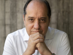 Foto de um homem branco, calvo, de cabelos escuros e curtos. Ele veste camisa social branca e relógio de pulso prateado. Sorri ligeiramente, com as mãos unidas sob o queixo e os cotovelos apoiados sobre uma mesa. O fundo é cinza e desfocado.