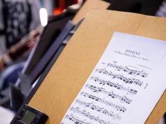 Foto da partitura da música Rondido. A página impressa está apoiada num suporte de madeira com um metrônomo apoiado no canto inferior esquerdo. Ao fundo, desfocada, do lado esquerdo, há uma pessoa tocando um instrumento de sopro.
