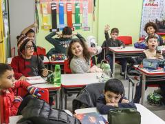 Foto de uma sala de aula. Há nove crianças sentadas em suas carteiras brancas e cadeiras vermelhas, enfileiradas em duplas. Algumas sorriem para a foto e outras olham em outras direções. Elas vestem roupas de frio. As paredes são verdes, com cartazes e materiais educativos e coloridos pendurados.