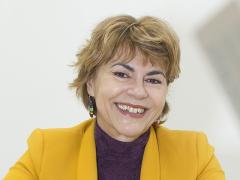 Foto de uma mulher branca, de cabelos loiros, curtos e lisos. Ela veste uma blusa roxa e um paletó amarelo. Ela está com as mãos uma sobre a outra e apoiadas sobre uma mesa. O fundo é branco.