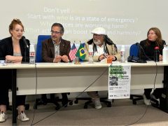 Foto de quatro pessoas sentadas em cadeiras azuis e diante de uma mesa comprida. Nas extremidades, duas mulheres brancas. A da esquerda é ruiva e fala ao microfone. A da direita tem cabelos castanhos claros e usa batom vermelho. Ao centro, dois homens. O primeiro é branco, usa óculos e tem cabelos grisalhos. O segundo é negro, usa uma boina branca, tem barba grisalha e dreads pretos. Sobre a mesa, repousam papéis, garrafas e bandeiras dos EUA e do Brasil. Atrás das pessoas, uma projeção em inglês na parede.