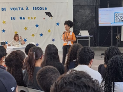 Foto de uma mulher negra falando ao microfone para um grupo de pessoas sentado à sua frente e de costas para a câmara. Ela está de pé, tem cabelos crespos que estão presos, usa uma camiseta laranja e uma calça preta. Atrás dela, uma mesa com duas mulheres e um painel branco onde está escrito “De volta à Escola”, “Eu na USP”, decorado com várias estrelas, em amarelo e azul. Atrás dela também há uma televisão que mostra um código QR. 