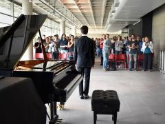 Foto de um pianista de pé diante de uma pequena plateia. O piano de cauda está em primeiro plano, à esquerda, e o pianista, usando terno, está ao centro e de costas para a câmera. Ao fundo, diversas pessoas, na maioria brancas, está de pé e batem palmas. As cadeiras do local são vermelhas e se destacam sobre o piso de concreto claro. 