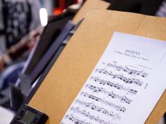 Foto de uma partitura impressa em uma folha de papel apoiada sobre um suporte de madeira. Ao lado dela, há um metrônomo digital e ao fundo há a silhueta de uma pessoa tocando um fagote.