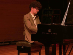 Foto de um homem negro de pele clara, sentado em um banco e tocando piano de cauda. Ele tem cabelos crespos, castanhos e curtos, veste um terno marrom com camisa branca e usa óculos de armação preto. O fundo é escuro.