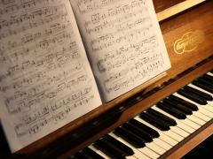 Foto de um piano com um livro de partituras aberto apoiado em seu suporte. O piano é de madeira, e parte do teclado com teclas pretas e brancas está visível. A partitura contém várias notas e anotações manuscritas. A luz ilumina a partitura e parte do piano.
