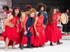 Foto de uma apresentação teatral em um palco. Um grupo de pessoas vestidas com roupas vermelhas está no centro da cena, olhando para a frente. Algumas estão em postura curvada e uma delas toca um tambor de mão. Ao fundo, outras pessoas, também vestidas de vermelho, seguram cadeiras brancas. O piso é claro e as paredes são escuras. 