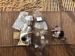 Foto de cima de duas mulheres sentadas em cadeiras sobre um piso de madeira. Ambas são brancas, de cabelos castanhos, mas uma delas aparenta ser mais jovem que a outra. Entre elas, há uma rede de pesca com papéis escritos dispostos sobre ela. Ao lado da mulher sentada à esquerda, um grande tecido claro coberto por escritos em azul.