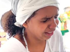 Foto de menina negra de olhos fechados e com expressão emocionada. Ela veste um turbante branco, tem cabelos castanhos escuros cacheados na altura do ombro e usa um fio de contas brancas e camiseta branca. O fundo é claro. 