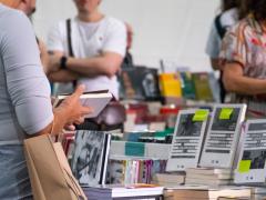 Foto de pessoa segurando livro em frente a uma bancada cheia de livros. Ao fundo estão livros e pessoas em desfoque.