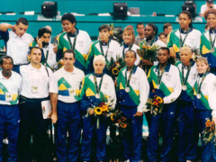  Foto da seleção brasileira feminina de basquete posando com medalhas de prata. Do lado esquerdo, o técnico e outros homens da comissão técnica. Os homens vestem calças pretas e azuis e camiseta branca com detalhes em verde, amarelo e azul. As mulheres vestem calça azul e agasalhos brancos com detalhes em verde, amarelo e azul. Uma pessoa à frente segura uma câmera de vídeo e veste bermuda bege e camiseta azul. Ao fundo está uma arquibancada e algumas pessoas.