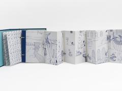 Foto de livro com capa azul e páginas brancas que se abrem em sanfona. Nas páginas, há diversos desenhos aparentemente feitos em caneta azul. O fundo é branco. 