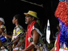 Foto de um grupo de pessoas negras de perfil, sendo que três estão em foco e seguram baquetas e tambores. Elas usam roupas coloridas de branco, amarelo, preto, vermelho e verde. Uma pessoa usa boina branca e outra um chapéu amarelo. O fundo é escuro e, no canto direito da imagem, há franjas vermelhas e azuis de papel celofane. 