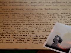  Em uma superfície, uma carta com escritos em letra cursiva e tinta preta e uma fotografia em preto e branco de uma mulher sobre a carta, no canto direito da imagem. A mulher tem cabelos curtos ondulados e sorri olhando na direção de algo que não aparece na foto. A carta é amarelada e a fotografia está com manchas amareladas também. 