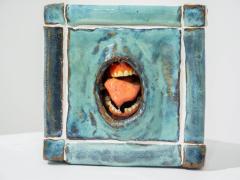 Foto de escultura em cerâmica de um cubo com uma boca aberta no centro. O cubo é azul e sua estrutura lembra pisos de cerâmica com rejunte branco. A boca é um buraco no centro do cubo e exibe língua, dentes e gengivas. 