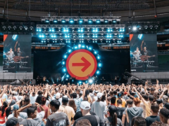 Foto de show musical com centenas de pessoas. A maioria do público é jovem e veste roupas claras. No palco, a banda e, ao fundo, um cenário com refletores e a imagem de uma seta vermelha em um círculo laranja, apontando para a direita. Acima, refletores com a cor azul. O palco é ladeado por telões que exibem imagens da banda.
