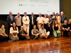 Foto de um grupo de 18 pessoas. Metade está em pé e a outra metade está sentada em um piso de madeira. São, em sua maioria, pessoas brancas que variam entre homens e mulheres, idosos e jovens. O fundo atrás deles é uma parede escura com uma tela grande com algumas informações projetadas, como a data, “28 de novembro de 2024”. 