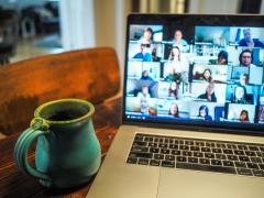 Foto de notebook sobre uma mesa. Na tela, há diversas imagens de pessoas em uma reunião online, ao lado do computador há uma caneca verde. 