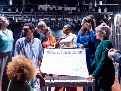 Foto de 9 pessoas, negras e brancas, em um palco de teatro.  Elas estão de pé, em torno de uma grande  folha de papel em que está impressa uma espécie de planta baixa . As pessoas vestem roupas coloridas. Ao fundo, há vários holofotes e uma parede preta.