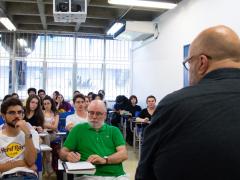 Foto de uma sala de aula. Um homem branco, calvo, usando oculos e blusa preta, está de costas. Um grupo de pessoas, sentadas em carteiras escolares, está a sua frente, e o observa. Ao fundo, janelas e uma parede branca.